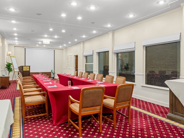 A conference room with a long U-shaped table, red tablecloths, beige chairs, white walls, large windows, and bright ceiling lights. Sentence ends here.