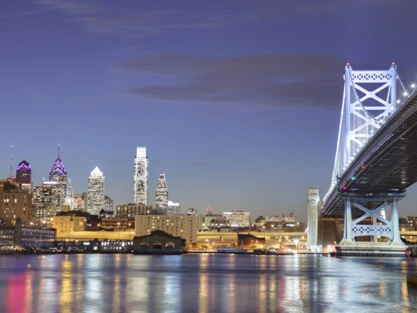 City skyline with illuminated skyscrapers and a lit-up bridge by the waterfront at dusk, reflecting on the water's surface.