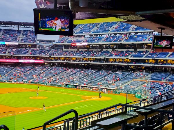 The image shows a baseball stadium interior with players on the field, some spectators, and screens displaying the game.