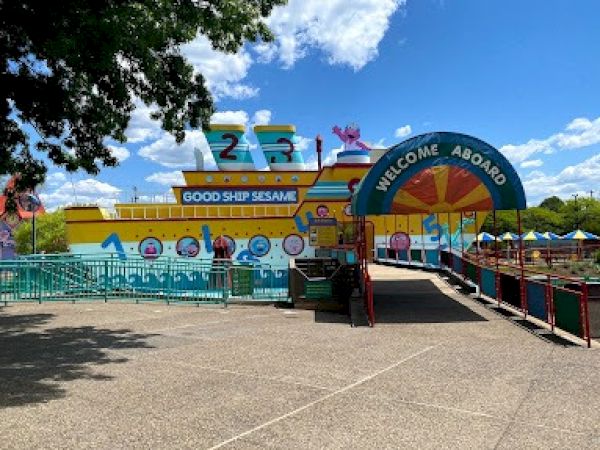 The image shows "Good Ship Sesame" at a theme park with colorful designs and a "Welcome Aboard" sign at the entrance.
