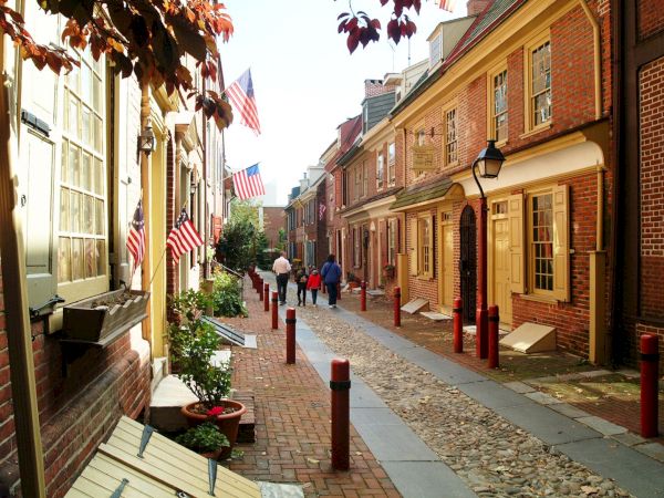 A narrow brick street lined with historic houses, adorned with American flags, and people walking down the path.