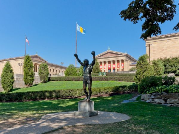 A statue of a boxer with raised arms stands before a classical building with flags and banners, surrounded by trees and neatly trimmed bushes.