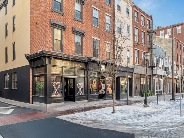 A row of brick storefronts on a snowy street, with large windows, pedestrians inside, and a tree-lined sidewalk.