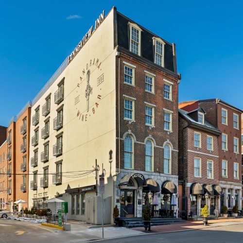 A street corner with a tan-brick building housing shops on the ground floor, modern windows above, and a clock or emblem on the tall facade, sunny blue sky.