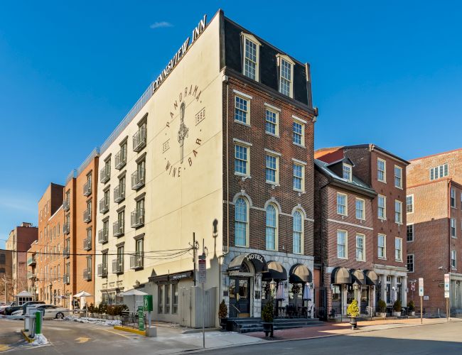 A street corner with a tan-brick building housing shops on the ground floor, modern windows above, and a clock or emblem on the tall facade, sunny blue sky.