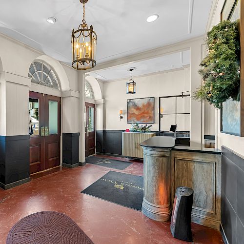A hotel lobby with polished timber doors, vintage lanterns, a reception desk, a luggage cart, arched windows, indoor plants, and warm lighting.