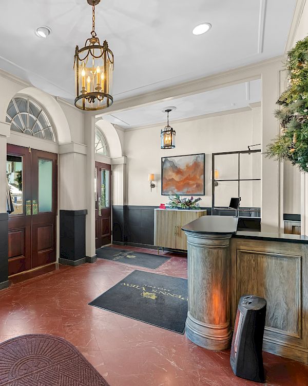 A hotel lobby with polished timber doors, vintage lanterns, a reception desk, a luggage cart, arched windows, indoor plants, and warm lighting.
