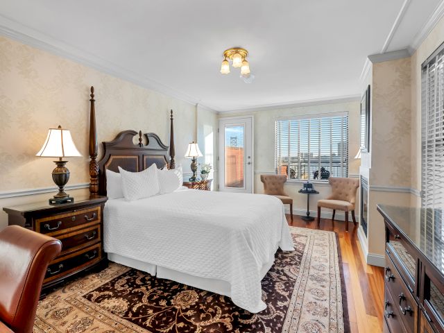 A luxurious hotel-style bedroom with a large wooden headboard bed, matching nightstands, two lamps, a rug, desk, chairs, and a windowed wall with blinds.