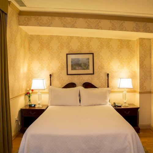 A cozy bedroom with a double bed, two lamps on bedside tables, patterned wallpaper, a picture on the wall, and curtains.