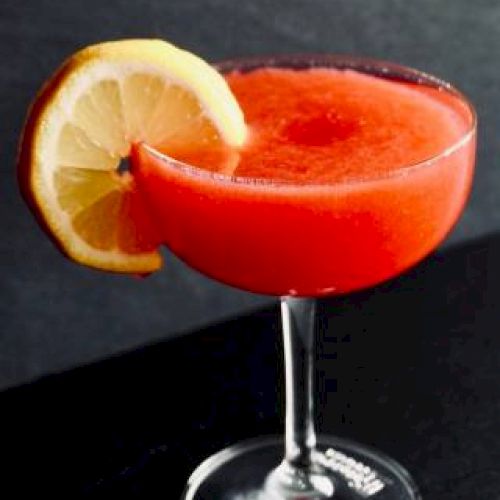 A red cocktail in a stemmed glass, garnished with a lemon slice, set against a dark background.