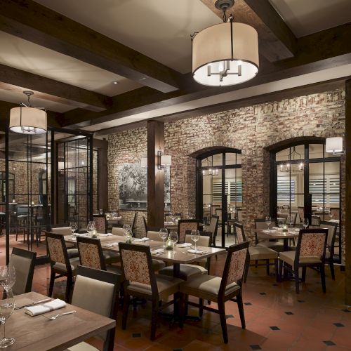 A cozy restaurant interior with brick walls, wooden beams, hanging lights, and neatly arranged tables and chairs creates a warm ambiance.