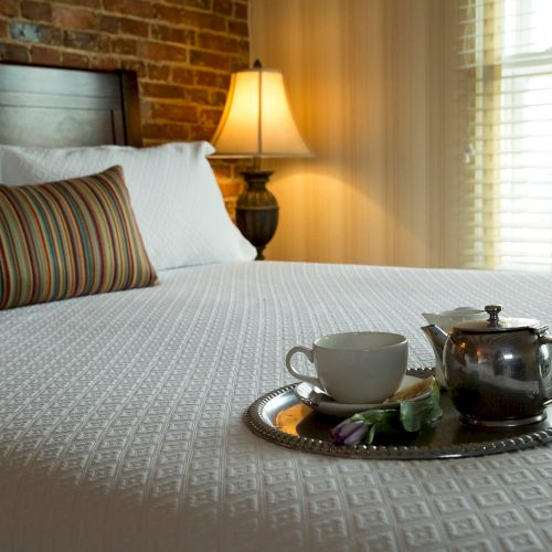 A cozy bedroom with a bed, striped pillow, lamp, and a tray holding a teapot, cup, and napkin, placed on a textured white bedspread.