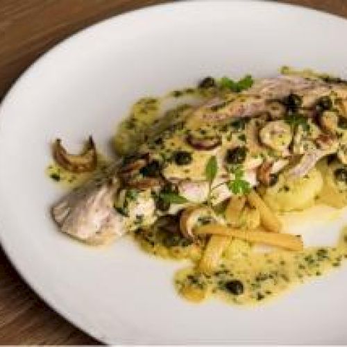 A plated fish dish with mushrooms, herbs, and a sauce on a white plate on a wooden surface.
