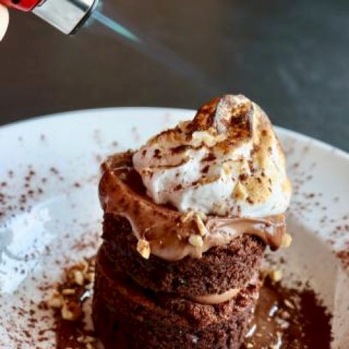 A chocolate dessert is topped with whipped cream, being torched, garnished with nuts and cocoa powder on a white plate.