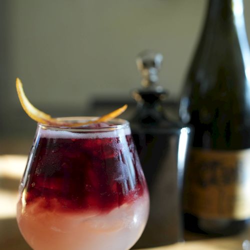 A cocktail with layered pink and red colors, garnished with a citrus twist, is placed on a table with bottles in the background.