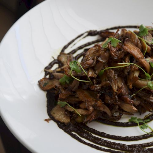 A dish featuring sautéed mushrooms, garnished with greens, arranged atop decorative dark sauce swirls on a white plate.