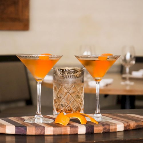 Two orange cocktails in martini glasses, an empty glass, and orange peels are placed on a wooden board.
