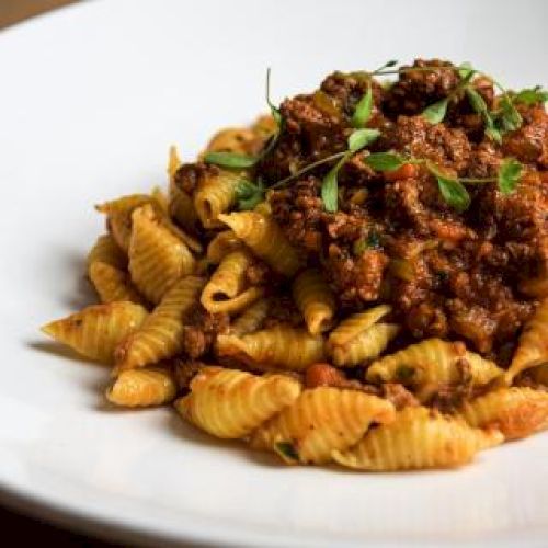 A white plate with pasta topped with a rich meat sauce and garnished with fresh herbs, placed on a wooden table.