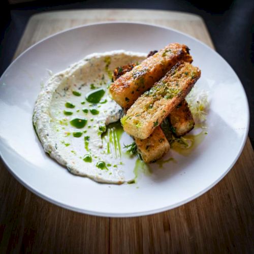 A white plate with breadsticks atop a creamy sauce garnished with herbs and drizzled with green oil.