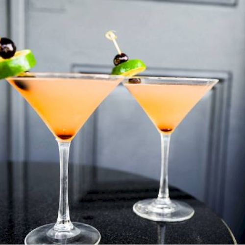 Two cocktails in martini glasses with lime slices and cherries garnish on a table in front of a dark background.
