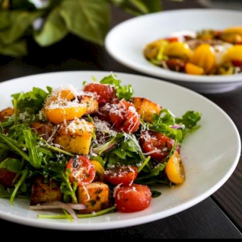 A plate of fresh salad with tomatoes, arugula, croutons, and grated cheese, with another similar plate in the background on a wooden table.
