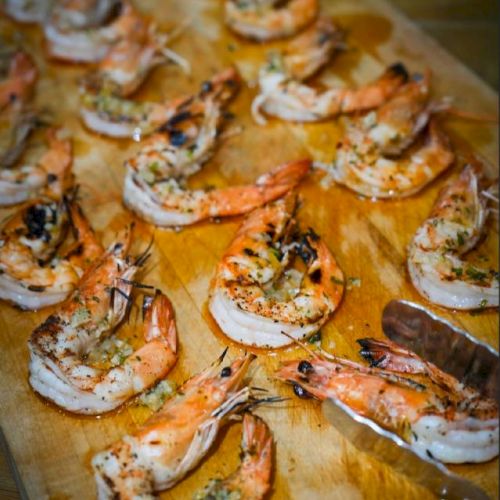 Grilled shrimp seasoned with herbs are arranged on a wooden board in a pattern.