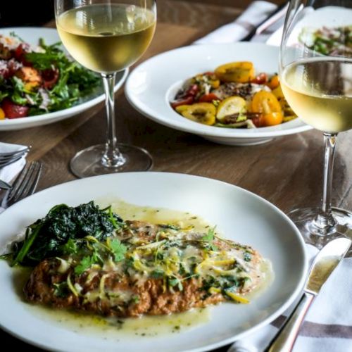 A table with plated gourmet dishes, featuring a main course and salads, paired with glasses of white wine.