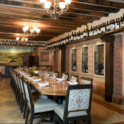 A long dining table set for guests in a rustic room with wine bottles on shelves and artwork on the walls, under warm lighting.