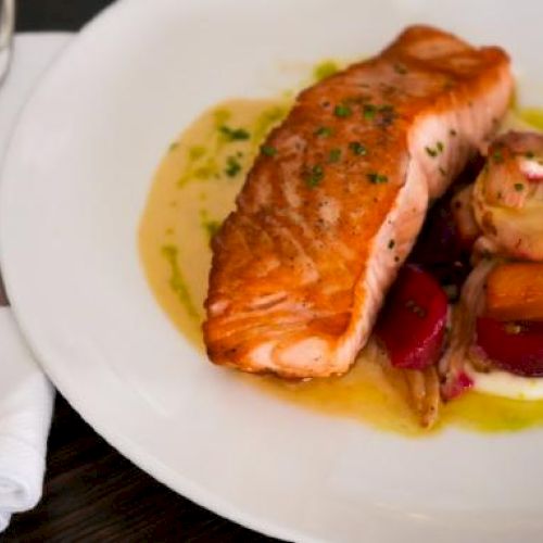 The image shows a cooked salmon fillet with a side of roasted vegetables on a white plate, accompanied by a fork and napkin.