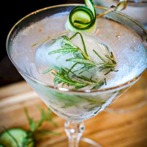 A cocktail with a large ice sphere, garnished with a cucumber twist and rosemary sprig, in a martini glass on a wooden surface.