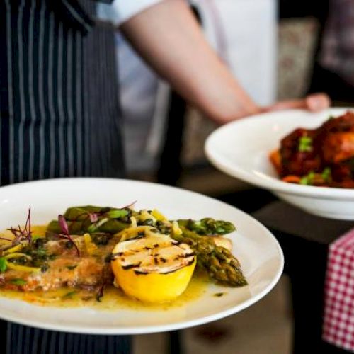 A person is holding two plates of food; one has a grilled lemon and vegetables, the other looks like a meat dish with sauce.