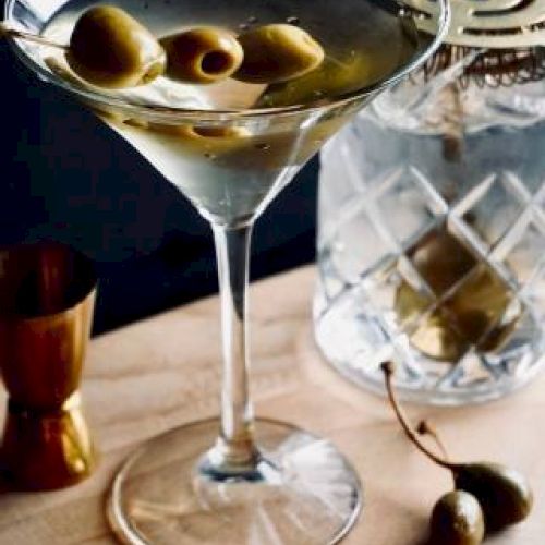 A martini garnished with olives sits on a wooden surface, accompanied by a jigger, a jar with a decorative lid, and yellow flowers.