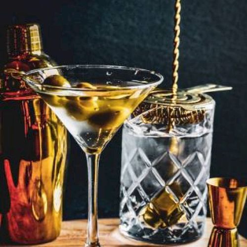 A cocktail setup with a martini glass, olives, cocktail shaker, mixing glass with spoon, and jigger displayed elegantly on a wooden surface.
