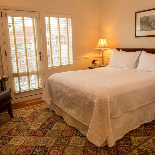 A cozy bedroom features a bed with white bedding, a chair, a lamp, and artwork on the wall, with light coming through window blinds.