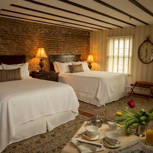 A cozy bedroom with two double beds, brick and striped walls, and a wooden tray with breakfast items and flowers on a table.