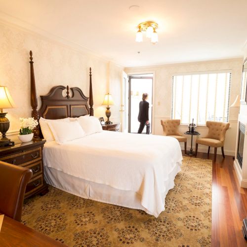 A cozy, well-lit bedroom with a large bed, lamps, chairs, a desk, and a person walking at the doorway, creating a warm and welcoming atmosphere.