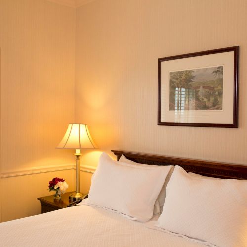 A cozy bedroom features a neatly made bed, bedside table with a lamp, a framed picture on the wall, and a window with blinds.