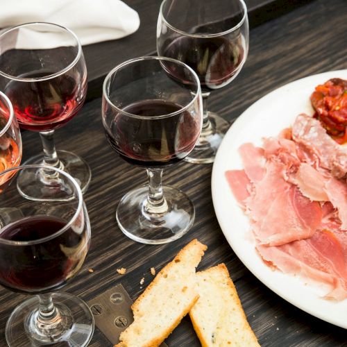A selection of five wine glasses, accompanied by a charcuterie platter with meats, cheeses, olives, and bread on a dark table.