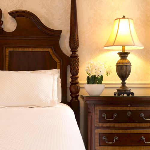 The image shows a cozy bedroom corner with a wooden bed, a lit lamp on a nightstand, and a potted plant for decoration.