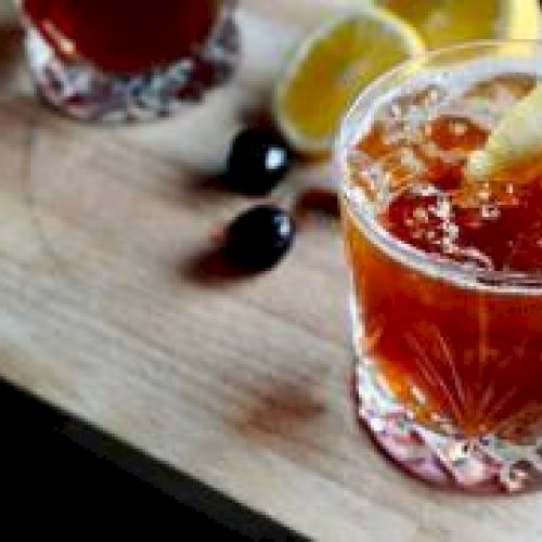 A cocktail garnished with a lemon twist and cherry sits on a wooden board, accompanied by lemon wedges and another drink in the background.