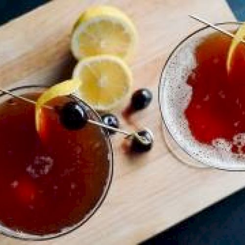 Two cocktails on a wooden board, garnished with lemon peel and cherries; sliced lemons are next to the drinks.