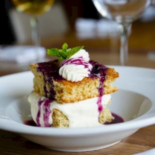 A dessert with layered cake, cream, and berry sauce, garnished with mint, served on a white plate, with wine glasses in the background.