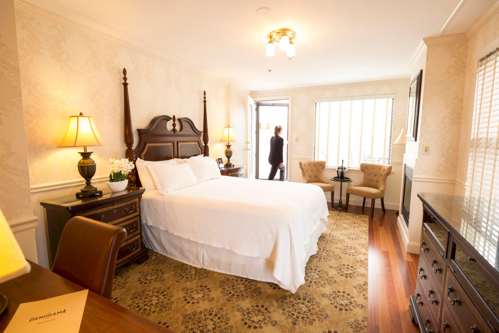 A cozy hotel room with a large bed, elegant furniture, and warm lighting. A person is standing near the window by a small table and chairs.