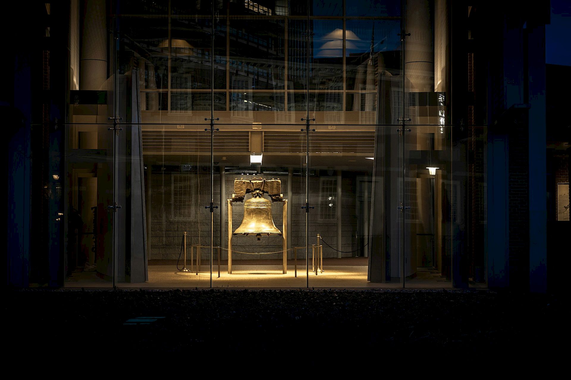 An illuminated bell displayed in a glass pavilion at night, set within a dimly lit building interior, surrounded by tall structures.