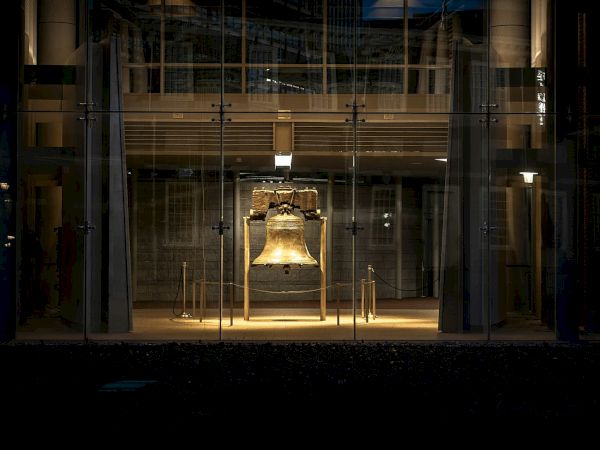 An illuminated bell displayed inside a glass-enclosed structure, situated in a dark environment.