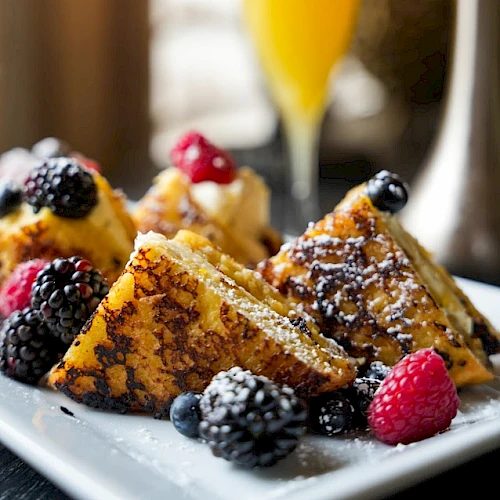 French toast topped with berries and powdered sugar on a plate, alongside an orange juice glass.
