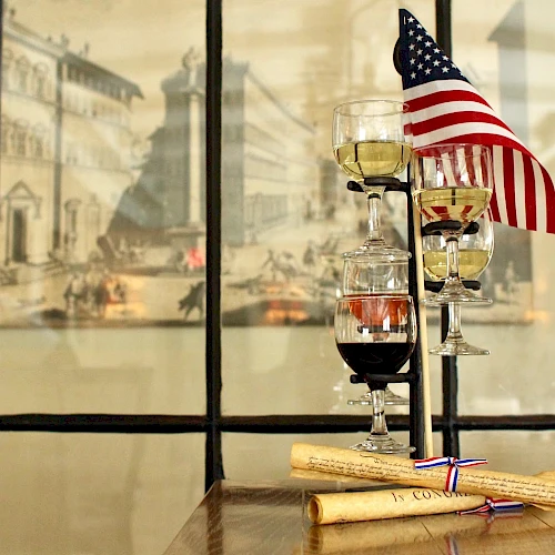 A tiered stand with three wine glasses and a U.S. flag, next to rolled parchment, in front of an artistic backdrop.