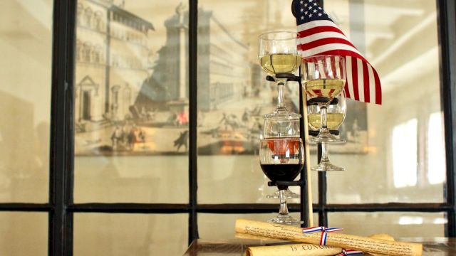 A tiered stand with three wine glasses and a U.S. flag, next to rolled parchment, in front of an artistic backdrop.