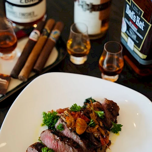 Sliced steak garnished with herbs and vegetables, whiskey bottles and glasses, and a tray with cigars are arranged on a table.