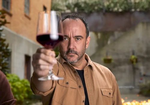 A man holds up a glass of red wine in an outdoor setting, focusing intently on it.